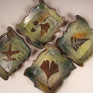 Op de afbeelding: Vier vierkante keramische schalen met gegolfde randen. Elke schaal heeft een ander bladontwerp in bruin op een groen-witte achtergrond.