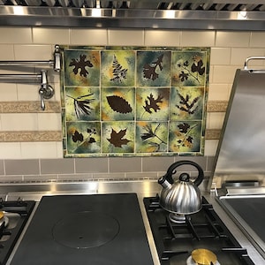 Puede incluir: Una encimera de cocina de acero inoxidable con un quemador negro y una tetera plateada en el lado derecho. El salpicadero está alicatado con un mosaico de hojas verdes y marrones.