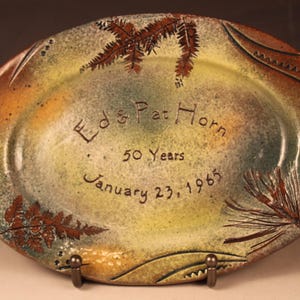 Puede incluir: Un plato ovalado de cerámica con un esmalte verde, marrón y amarillo. El plato está decorado con diseños de helechos y pinos. Tiene la inscripción "Ed & Pat Horn 50 Years January 23, 1965".