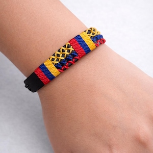 Może przedstawiać: Tkana bransoletka z czarnym paskiem i kolorowym centralnym wzorem. Wzór zawiera żółte, czerwone i niebieskie nici w geometryczny wzór. Bransoletka jest noszona na nadgarstku.