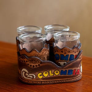 May include: A set of four clear glass shot glasses in a brown leather holder. The holder is decorated with the word "COLOMBIA" in yellow, blue, and red lettering. The holder has a decorative wavy edge.