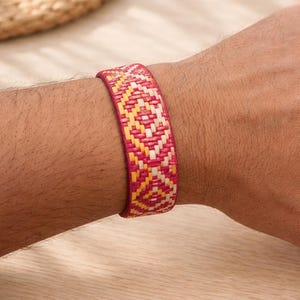 May include: A woven bracelet with a geometric pattern in shades of pink, yellow, and white. The bracelet is approximately 1.5 inches wide and is worn on a wrist. The design features a repeating diamond pattern.