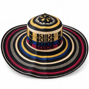 May include: A wide-brimmed woven hat featuring a cylindrical crown with a geometric star pattern on top. The hat displays concentric bands of black, cream, yellow, blue, and red, with intricate woven patterns around the crown and brim edges.