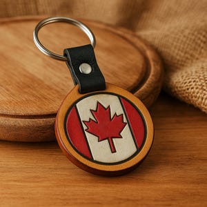 Puede incluir: Un llavero redondo de cuero con un diseño de bandera canadiense. El llavero es marrón con una hoja de arce roja sobre un fondo blanco. El llavero tiene una correa de cuero negro y un anillo de metal plateado.