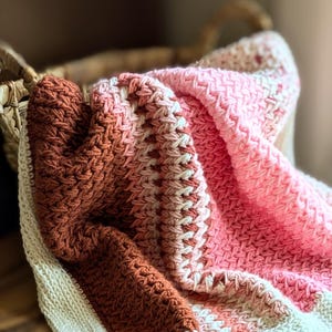 May include: A crocheted baby blanket with a pink and brown ombre pattern. The blanket is folded and lying on a wooden surface with two wooden baby toys.