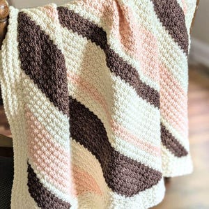 May include: A handmade crochet blanket draped over a wooden chair. The blanket features a textured stitch pattern with diagonal stripes in cream, light pink, and dark brown. The blanket is soft and cozy, perfect for snuggling up.