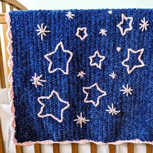 Peut inclure: Une couverture pour bébé bleue avec une bordure en crochet rose et des étoiles brodées roses.