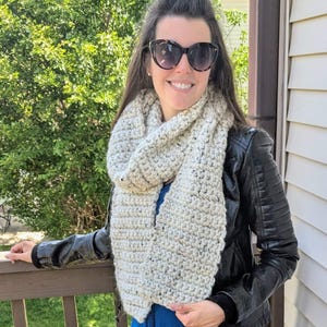 May include: A light beige, chunky-knit scarf is draped over a person's shoulders. The scarf is made of a thick yarn with a textured appearance. The person is wearing a black leather jacket and a blue dress. Sunglasses are worn.