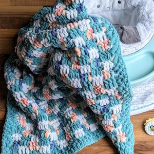 May include: A teal, white, pink, and blue crocheted baby blanket with a textured, fluffy look. The blanket is draped over a baby bouncer.
