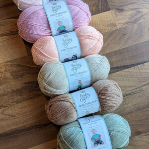 May include: An assortment of Baby Bee Sweet Delight yarn in various pastel colors, including pink, peach, and green. Each skein has a label with a baby illustration. The yarn is arranged on a wooden surface.