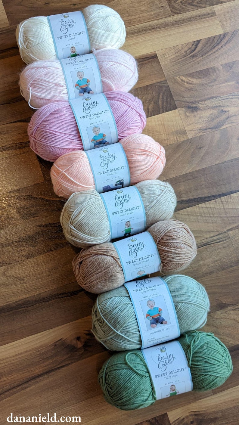 May include: An assortment of Baby Bee Sweet Delight yarn in various pastel colors, including pink, peach, and green. Each skein has a label with a baby illustration. The yarn is arranged on a wooden surface.
