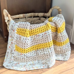 May include: A hand-crocheted blanket featuring horizontal stripes of mustard yellow and speckled beige yarn. The textured fabric displays a repeating stitch pattern with a speckled border in white, beige, and orange tones, draped over a woven basket.