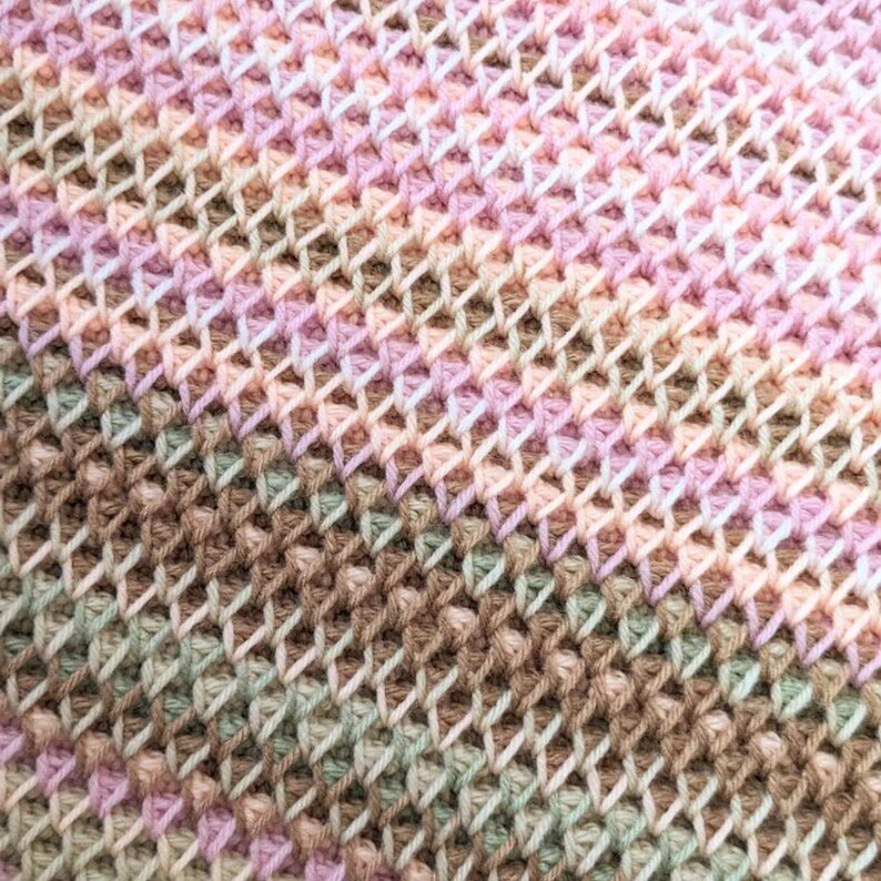 May include: Close-up of a crocheted fabric with a textured, open weave. The color palette includes soft hues of pink, peach, brown, and green, creating a gradient effect. The stitches form a repeating pattern.