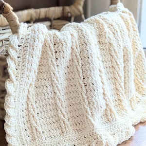 May include: A cream-colored crocheted blanket featuring a textured cable stitch pattern with vertical columns of twisted yarn. The blanket has a decorative scalloped border along the edge and includes the text dananield.com printed at the bottom.