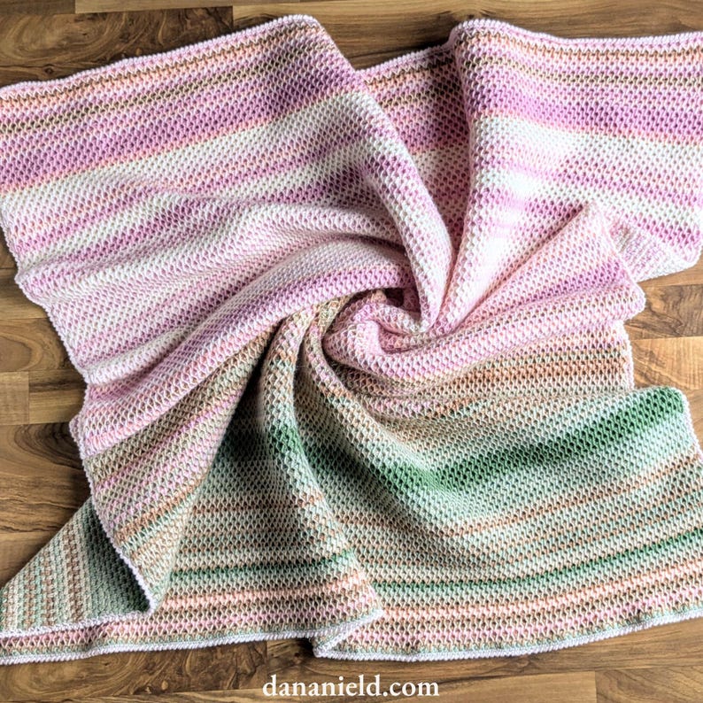 May include: A knitted blanket with a striped pattern in shades of pink, white, green, and brown. The blanket is folded and draped, showcasing the texture and color variations. The blanket is made of soft material.