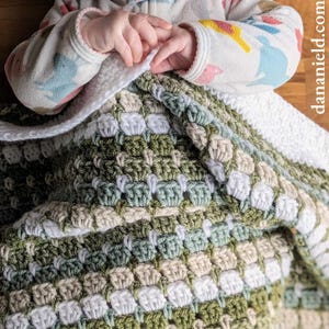 May include: A crocheted baby blanket in a green, white and beige colour scheme. The blanket is made with a textured stitch pattern.