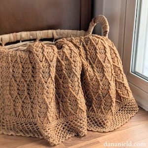 May include: A tan crocheted blanket with a diamond pattern is draped over a woven basket. The blanket's intricate design and warm color create a cozy, inviting feel. The basket has a curved handle.