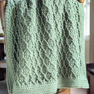 May include: A sage green crocheted blanket featuring a raised diamond cable stitch pattern. The textile has a thick, textured appearance with a wide, flat-stitched border along the bottom edge, showcasing a consistent yarn colour throughout.