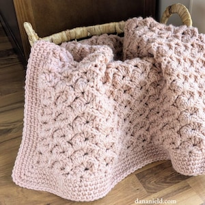 May include: A blush pink crocheted blanket with a textured pattern, folded in a wicker basket. The blanket's intricate stitchwork creates a visually appealing design, suitable for home decor or as a cosy accessory.
