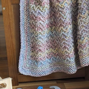 May include: A multi-colored crocheted blanket with a chevron pattern hangs over a wooden crib. The blanket is made with a variety of shades of pink, blue, and green yarn.