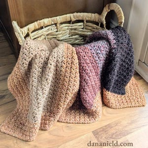 May include: A collection of handmade crochet blankets in a woven basket. The blankets display a gradient of colors, transitioning from light tan to shades of pink and deep charcoal. The blankets are folded and draped, showcasing their textured stitch.