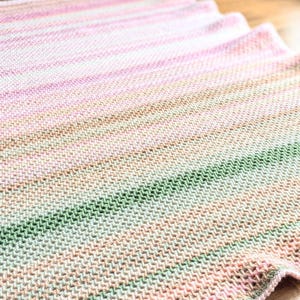 May include: A hand-knitted blanket in shades of pink, green, and tan, with a textured stitch pattern. The blanket is lying on a wooden floor, with the edge of the blanket slightly curved. The blanket is a soft, cozy home decor item.
