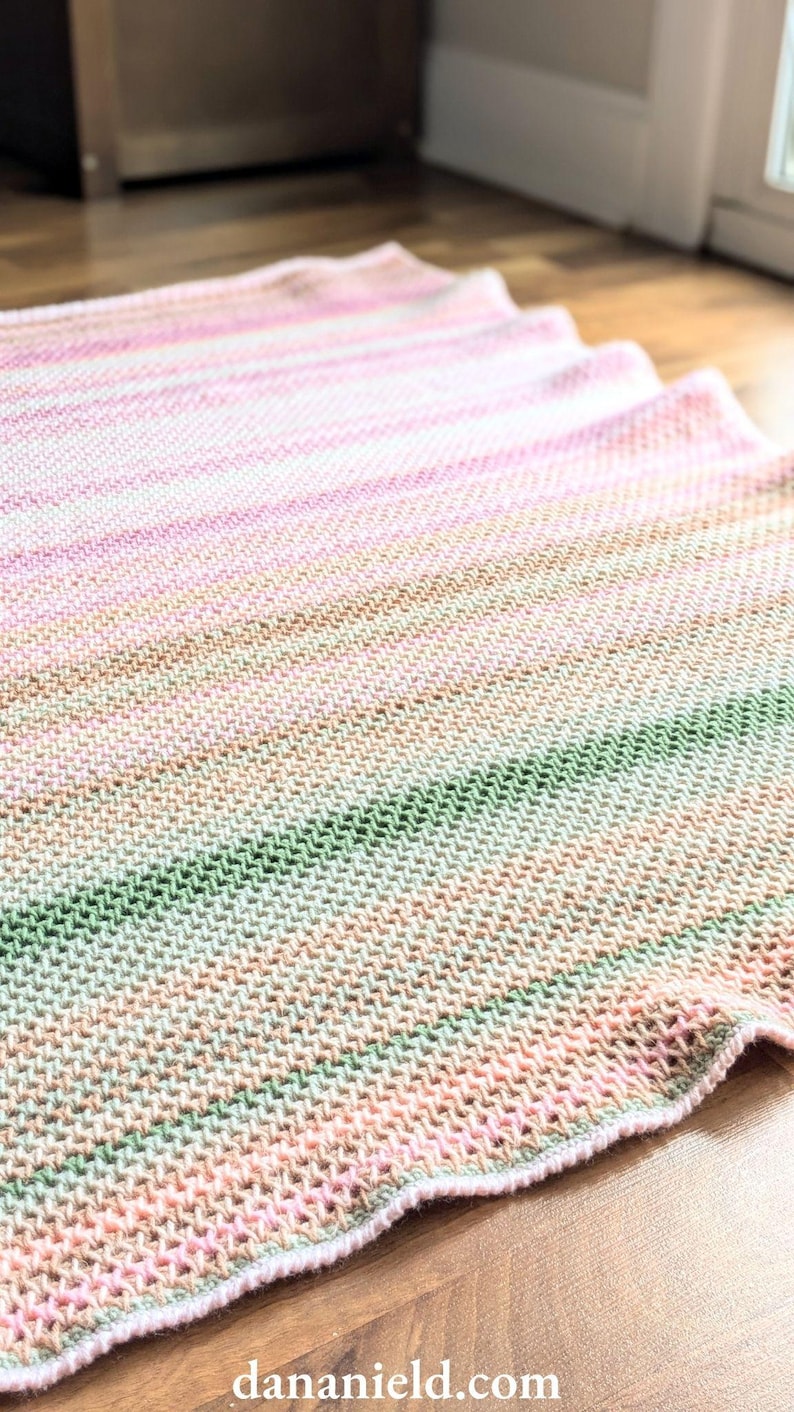 May include: A hand-knitted blanket in shades of pink, green, and tan, with a textured stitch pattern. The blanket is lying on a wooden floor, with the edge of the blanket slightly curved. The blanket is a soft, cozy home decor item.