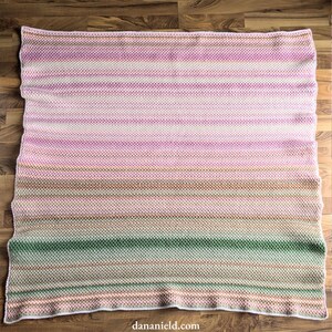 May include: A handmade, rectangular blanket with a horizontal striped pattern. The color palette transitions from pink to green, with shades of white, brown, and peach. The blanket is made of a textured knit fabric.