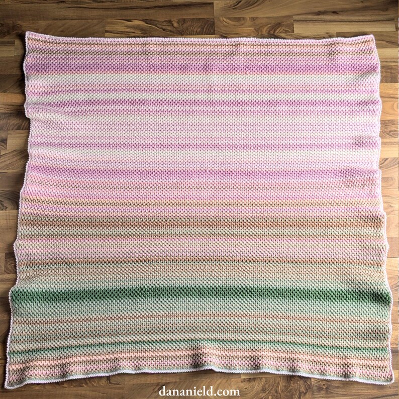 May include: A handmade, rectangular blanket with a horizontal striped pattern. The color palette transitions from pink to green, with shades of white, brown, and peach. The blanket is made of a textured knit fabric.