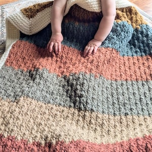 May include: A handmade, textured baby blanket with horizontal stripes in shades of peach, gray, beige, and blue. The blanket is made of a soft, chunky yarn. The baby's hands and feet are visible.