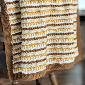 May include: A handmade crochet blanket draped over a wooden chair. The blanket features horizontal stripes in shades of brown, cream, and gold. The border is a solid brown, and the blanket has a textured appearance.