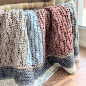 May include: A crochet blanket featuring vertical panels in beige, light blue, dusty rose, and grey, with a textured cable stitch pattern. The bottom edge has horizontal stripes in dark grey, light grey, and cream, with the text dananield.com.