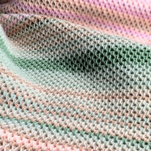 May include: Close-up of a soft, hand-knitted blanket. The blanket features a textured, open weave design with a gradient of colors, including pink, green, and beige. The blanket is folded, showing the intricate stitch pattern and color variations.