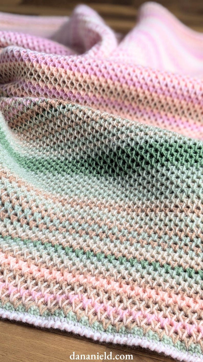 May include: Close-up of a soft, hand-knitted blanket. The blanket features a textured, open weave design with a gradient of colors, including pink, green, and beige. The blanket is folded, showing the intricate stitch pattern and color variations.