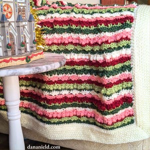 Puede incluir: Una manta de crochet a rayas con un borde blanco. La manta está hecha con diferentes tonos de rojo, verde, rosa y blanco. La manta está drapeada sobre un sofá.