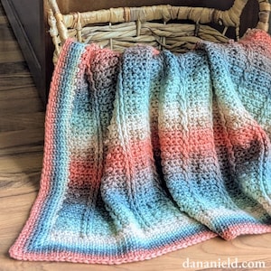 May include: A handmade, multi-colored crochet blanket with a gradient of colors including coral, blue, and white. The blanket is folded over a wicker basket, showcasing its textured stitch pattern and soft, inviting appearance. The blanket's edge is finished with a coral border.