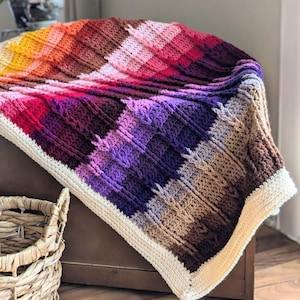 May include: A colourful crocheted blanket with a striped pattern in shades of orange, red, purple, brown, and white. The blanket is folded and draped over a wooden surface.