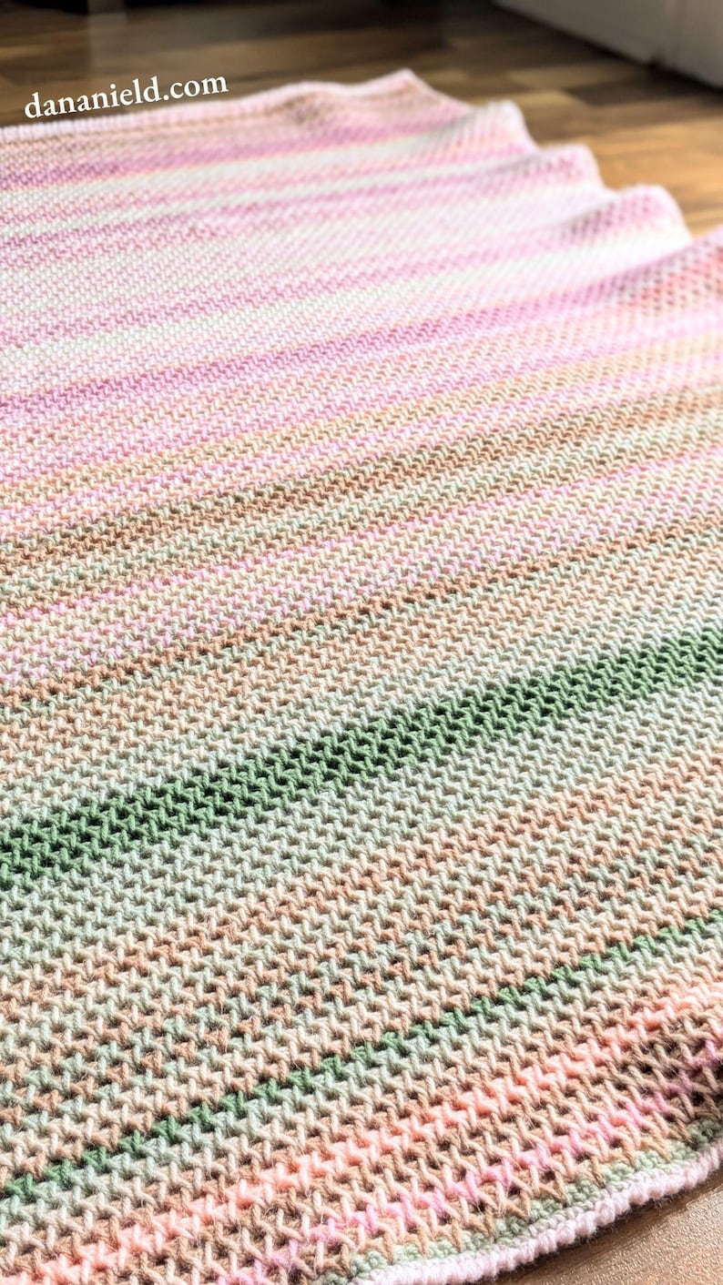 May include: A hand-knitted blanket with a gradient of colors, including pink, green, and tan. The blanket has a textured appearance and is made of soft yarn. The website address dananield.com is visible in the image.