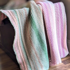 May include: A soft, textured blanket in shades of pink, green, and cream. The blanket is draped over a dark brown surface, with the textured pattern clearly visible. The color gradient creates a gentle, inviting aesthetic.