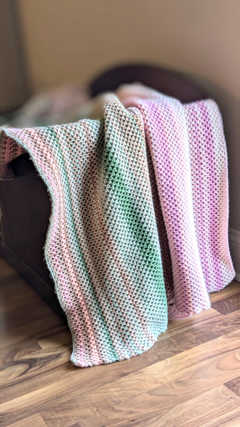 May include: A soft, textured blanket in shades of pink, green, and cream. The blanket is draped over a dark brown surface, with the textured pattern clearly visible. The color gradient creates a gentle, inviting aesthetic.