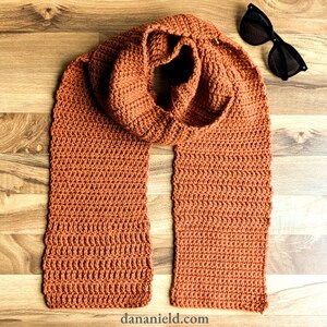 May include: A long, rust-coloured crocheted scarf with a simple stitch pattern. The scarf is folded in half and laid flat on a wooden surface.