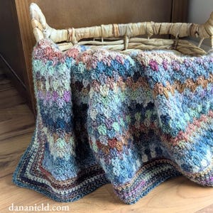 May include: A handmade crochet blanket in a wicker basket. The blanket features a textured stitch pattern in shades of blue, green, purple, and brown. The blanket is folded over the edge of the basket, showcasing its intricate design and color palette.