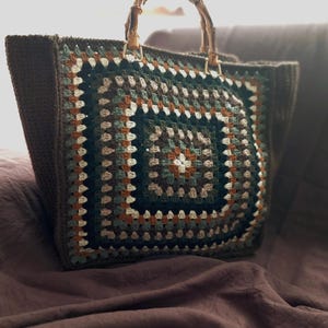 May include: A handmade crochet tote bag with a square pattern in shades of brown, green, and cream. The bag features bamboo handles and a sturdy, dark brown base. The crochet design is a classic granny square pattern.