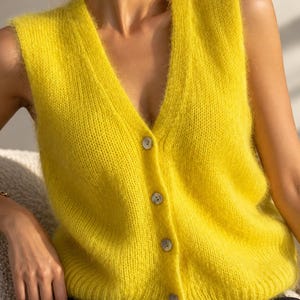 Puede incluir: Un chaleco de punto sin mangas en color amarillo brillante con un escote en V profundo y cierre frontal de cuatro botones claros. La prenda está confeccionada con un hilo de textura suave y esponjosa e incluye un dobladillo acanalado.