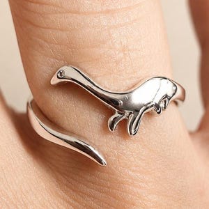 Könnte beinhalten: Ein silberfarbener offener Ring mit einem polierten Dinosaurier-Design, das sich um den Finger legt. Der Ring zeigt die Silhouette eines langhalsigen Dinosauriers mit einer glatten, reflektierenden Metalloberfläche, die ein offenes Band bildet.