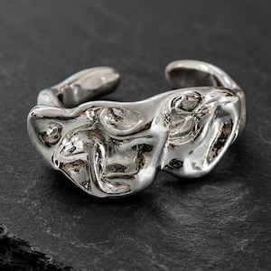 Op de afbeelding: Een zilverkleurige ring met een open band en een zeer onregelmatige, gesmolten oppervlaktestructuur. Het metaal heeft een gepolijste, reflecterende afwerking met diepe inkepingen en verhoogde randen over de gehele buitenomtrek.