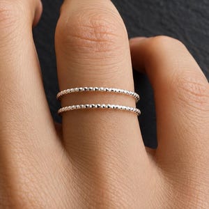 Könnte beinhalten: Zwei dünne, silberfarbene Ringe mit einer texturierten, perlenartigen Oberfläche. Jeder Ring weist ein feines, sich wiederholendes Muster aus kleinen, erhabenen Metallpunkten entlang des gesamten Umfangs auf, was für ein subtil reflektierendes Aussehen sorgt.