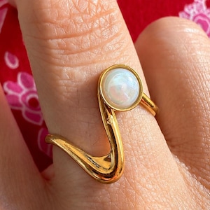 Könnte beinhalten: Ein goldfarbener Ring mit einer glatten, geschwungenen S-förmigen Ringschiene, die sich um den Finger legt. Der Ring ist mit einem einzelnen, runden, schimmernden weißen Cabochon-Stein besetzt, der in einer runden Fassung oben sitzt.