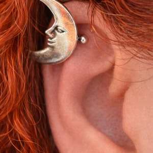 Könnte beinhalten: Silberfarbener Ohrschmuck in Form eines Halbmonds mit detailliertem Gesichtsdesign, getragen am Ohr. Der Ohrschmuck wird mit einem kleinen, runden Kristallohrstecker kombiniert. Rot-oranges Haar umrahmt das Ohr und erzeugt einen auffälligen Kontrast.