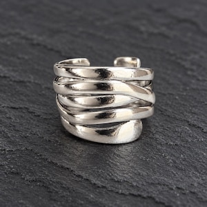 Pode incluir: Um anel de tom prateado com um design de banda aberta de várias camadas. O anel é composto por várias bandas lisas e sobrepostas que envolvem o dedo, criando uma aparência larga, contínua e fluida com um acabamento metálico polido.